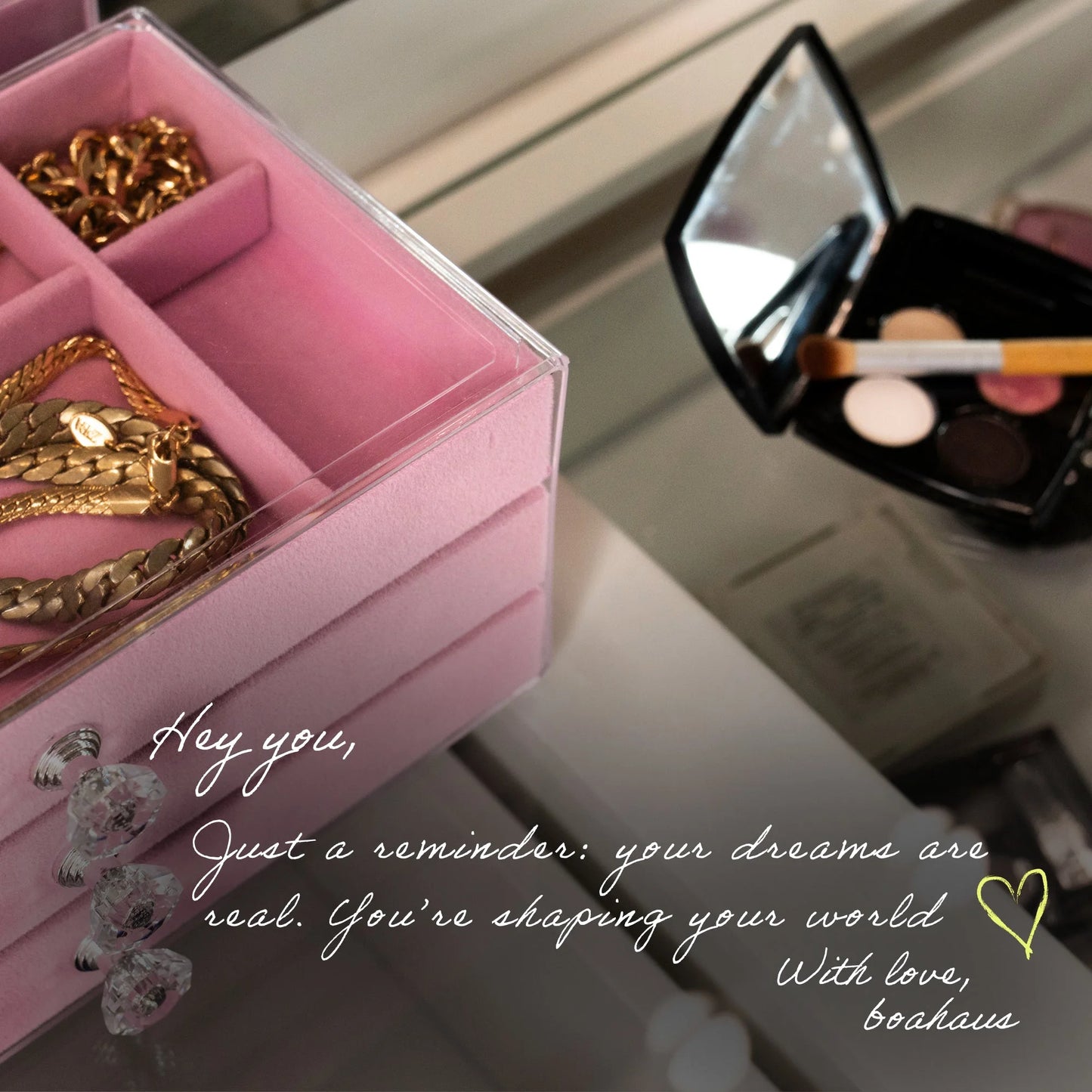 Pink jewelry box with gold chains, makeup compact with brushes on a reflective surface.